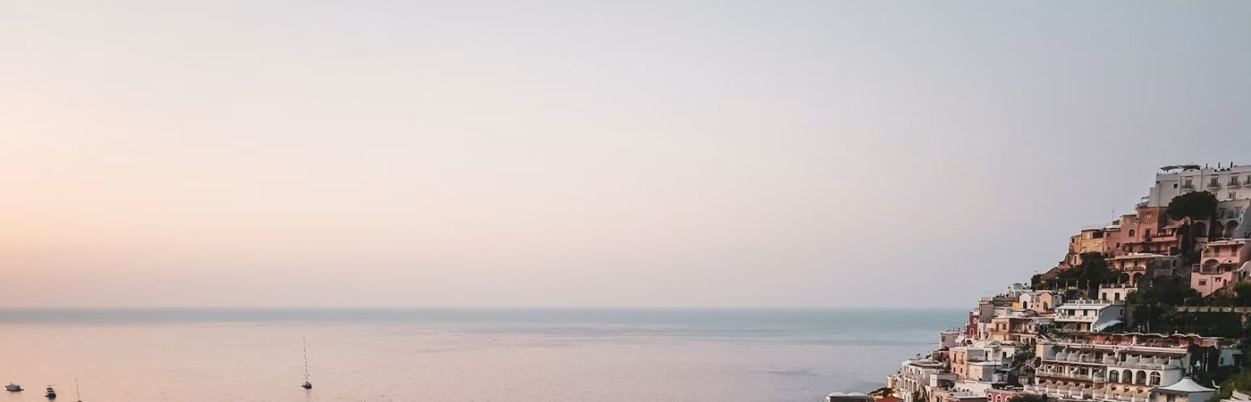 A pink-and-white coastal town on a cliffside above the sea at dusk, boats scattered across the water